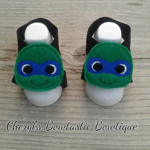 May include: A pair of black and green baby sandals with a Teenage Mutant Ninja Turtles character design. The sandals have a velcro strap closure and are made of soft fabric.