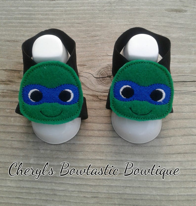 May include: A pair of black and green baby sandals with a Teenage Mutant Ninja Turtles character design. The sandals have a velcro strap closure and are made of soft fabric.