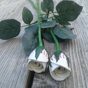 May include: Two white leather roses with red stitching, resembling baseballs, are displayed on a wooden surface with green leaves.