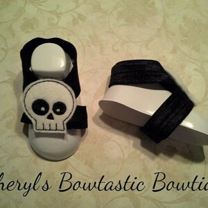 May include: A pair of black and white baby sandals with a skull design. The sandals are made of fabric and have a white sole.