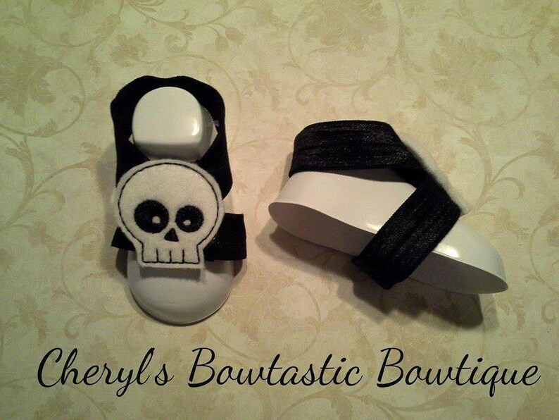 May include: A pair of black and white baby sandals with a skull design. The sandals are made of fabric and have a white sole.