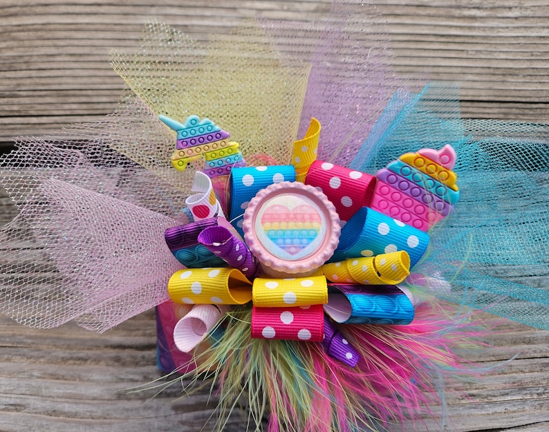 May include: A colourful hair bow featuring a rainbow heart, polka dot ribbon and a unicorn charm. The bow is made with tulle and feathers.