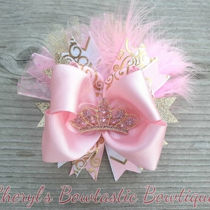 May include: A pink hair bow with a gold tiara, feathers, and glitter accents. The bow features a large pink satin ribbon, gold patterned ribbon, and a pink tulle overlay. The text "Cheryl's Bowtastic Boutique" is at the bottom.