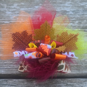 May include: A fall-themed hair bow featuring orange, red, and yellow tulle, with decorative leaves and ribbons. The bow includes a variety of textures and colors, including brown, orange, and white ribbons, and burgundy feathers.