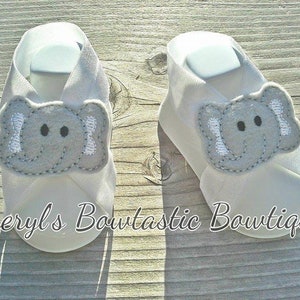 May include: A pair of white baby booties with an embroidered gray elephant design. The booties have a criss-cross strap design and are made of soft fabric.
