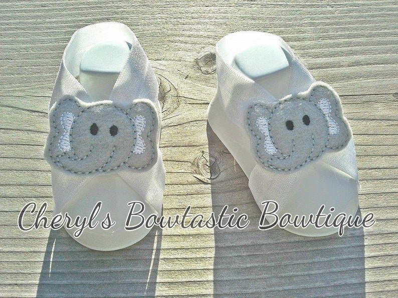 May include: A pair of white baby booties with an embroidered gray elephant design. The booties have a criss-cross strap design and are made of soft fabric.