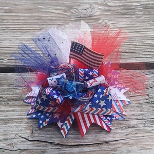 May include: A hair bow featuring red, white, and blue ribbons with star and stripe patterns, layered with red, white, and blue tulle. A small American flag is attached to the centre, along with a blue star-shaped plastic accent piece.