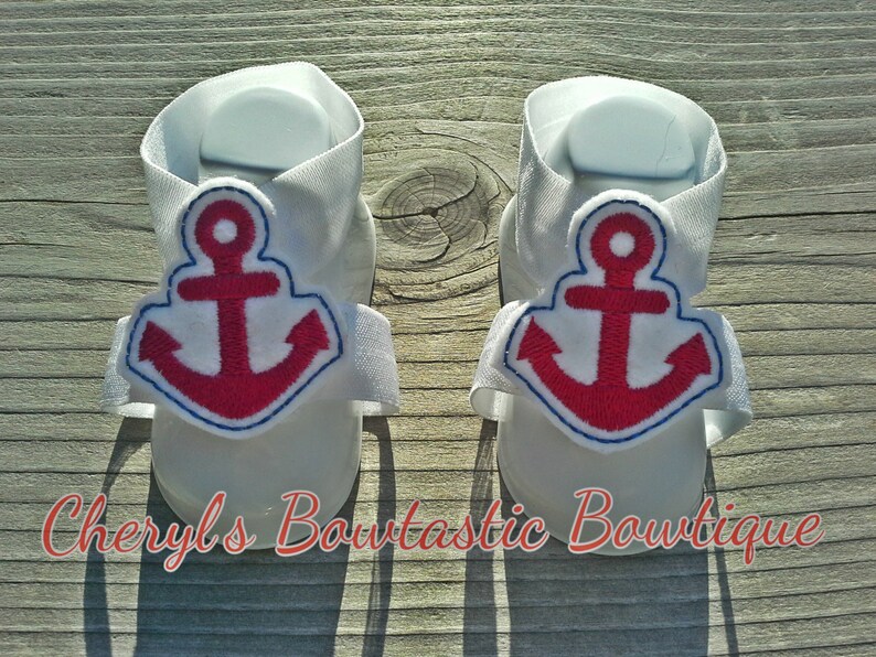 May include: A pair of white baby sandals with red anchor embroidery. The sandals have adjustable straps and are perfect for summer.