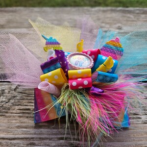 May include: A colourful hair bow featuring pink, yellow, blue and purple ribbon. The bow includes a pop-it toy and a pink button with the text "Pop It" on it. The bow is decorated with white tulle and a pink and white polka dot ribbon.
