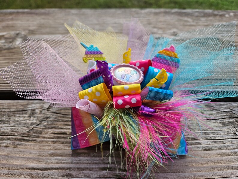May include: A colourful hair bow featuring pink, yellow, blue and purple ribbon. The bow includes a pop-it toy and a pink button with the text "Pop It" on it. The bow is decorated with white tulle and a pink and white polka dot ribbon.