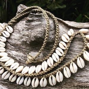 May include: A necklace made of white cowrie shells strung on a brown cord. The necklace is displayed on a piece of weathered wood.