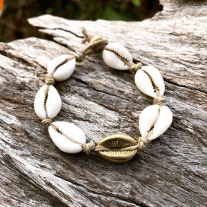 Peut inclure: Un bracelet en coquillages blancs avec un charme doré, attaché avec un cordon en fibre naturelle. Le bracelet est sur une surface en bois.