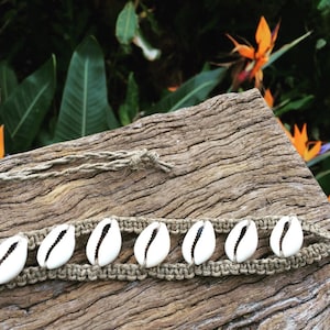 Op de afbeelding: Een bruin hennep armband met witte kauri schelpen. De armband is vastgemaakt met een knoop en heeft een lange kwast.
