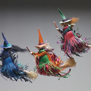 May include: Three handmade witches on broomsticks, each with a different colored dress and hat. The witches are made of straw and fabric and have long, flowing hair. The broomsticks are also made of straw and have a natural, rustic look.