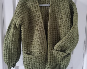 Pode incluir: Um casaco de tricô verde com mangas compridas e bolsos. O casaco está pendurado em um cabide.