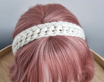 May include: A white crocheted headband with a textured design. The headband is worn on a mannequin with pink hair.