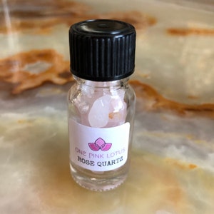 May include: A small glass bottle filled with rose quartz crystals. The bottle has a black lid and a white label that reads "One Pink Lotus Rose Quartz".