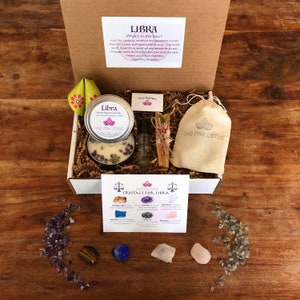 May include: A white gift box filled with a Libra themed gift set. The box contains a white candle with amethyst crystals, a small bottle of oil, a sage smudge stick, a small pouch, and a card with information about crystals for Libra. The box is sitting on a wooden surface.