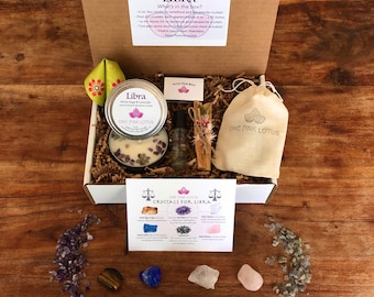 May include: A white gift box filled with a Libra themed gift set. The box contains a white candle with amethyst crystals, a small bottle of oil, a sage smudge stick, a small pouch, and a card with information about crystals for Libra. The box is sitting on a wooden surface.