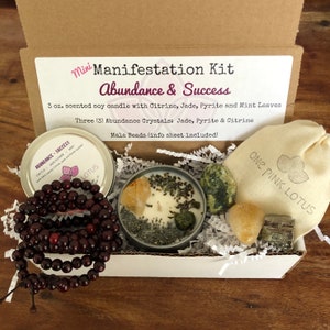 May include: A white box filled with brown paper. The box contains a 3 oz. soy candle with citrine, jade, pyrite, and mint leaves. There are three abundance crystals: jade, pyrite, and citrine. A mala bead necklace is also included. The box has a label that reads "Mini Manifestation Kit Abundance & Success".