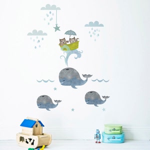 May include: A set of three blue whale wall decals with a smaller whale on each side of a larger whale. The larger whale has a pair of cats in a yellow boat with an umbrella above them. The cats are wearing blue and green hats. There are clouds and stars in the background.