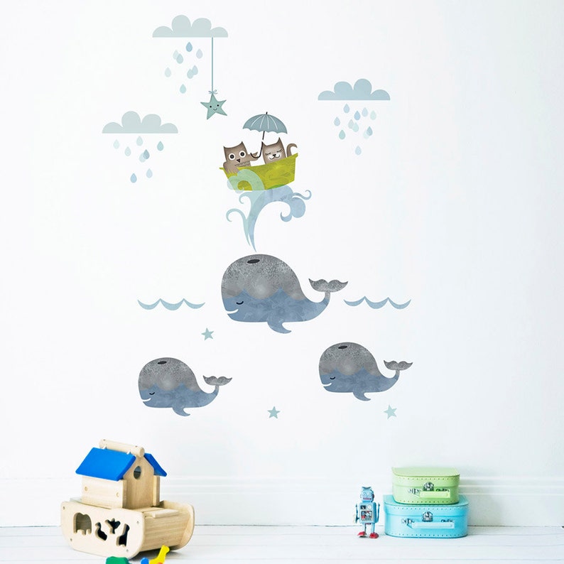 May include: A set of three blue whale wall decals with a smaller whale on each side of a larger whale. The larger whale has a pair of cats in a yellow boat with an umbrella above them. The cats are wearing blue and green hats. There are clouds and stars in the background.