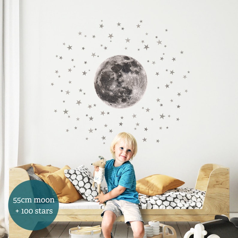Puede incluir: Un ni&ntilde;o se sienta en una cama con una gran luna gris y 100 peque&ntilde;as estrellas grises en pegatinas de pared. La luna tiene un di&aacute;metro de 55 cm. El texto "55cm moon + 100 stars" es visible en la imagen.