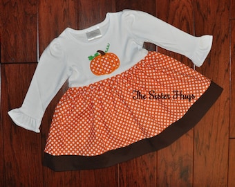 May include: A long-sleeved white dress featuring a pumpkin appliqué on the bodice and an orange skirt with white polka dots and a solid brown hem. The text The Sister Hugs is printed on the skirt. The sleeves have ruffled cuffs.