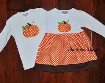 May include: A white long-sleeve shirt and a white long-sleeve dress, both featuring an embroidered orange pumpkin with white polka dots. The dress has an orange polka-dot skirt with a brown hem. The text The Sister Hugs is visible.