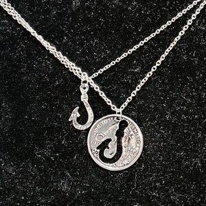 May include: A silver necklace with a hook charm and a silver coin pendant with a mermaid silhouette cut out. The coin pendant has the words "United States of America" and "Liberty" inscribed on it.