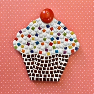 May include: A mosaic cupcake with a red cherry on top. The cupcake is made of small, colorful tiles and is decorated with white frosting and colorful sprinkles.