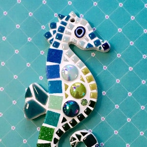 May include: A mosaic seahorse in shades of blue, green, and white. The seahorse is decorated with square tiles, round gems, and buttons. The background is a light blue surface with a floral pattern.