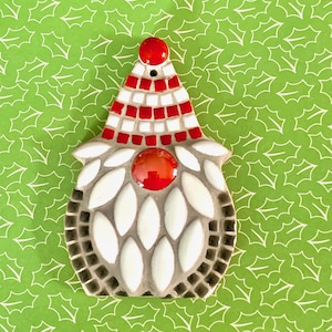 May include: A white and red mosaic gnome ornament with a red hat and a white beard. The gnome is sitting on a green background with a holly leaf pattern.