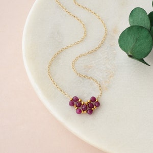 May include: A delicate gold chain necklace featuring a small, V-shaped pendant composed of several faceted, deep magenta-colored gemstone beads interspersed with tiny, round, polished gold-tone metal spacer beads in a cluster arrangement.