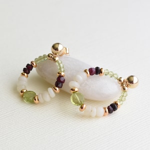 May include: A pair of gold hoop earrings featuring a mix of green, white and red beads. The earrings are made with a variety of gemstones, including peridot, white opal and ruby.