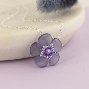 May include: A delicate silver chain necklace with a purple flower pendant. The flower is made of translucent plastic and has a beaded center.