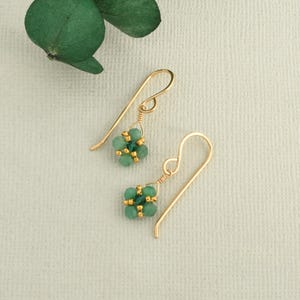 May include: A pair of gold earrings with green gemstone accents. The earrings are made with small, round beads that are clustered together to form a flower shape.