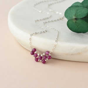 May include: A delicate silver chain necklace featuring a cluster of small, faceted, deep pink ruby-colored beads interspersed with tiny silver metallic spheres, creating a compact, multi-layered pendant arrangement on a thin, polished silver link chain.