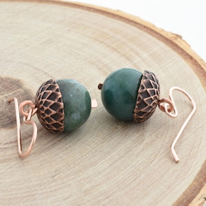 May include: A pair of acorn-shaped earrings. Each earring features a dark green, round bead paired with a copper-colored, textured acorn cap. The earrings have copper-colored ear wires. The earrings are displayed on a light brown, wooden surface.