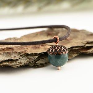 May include: A handcrafted necklace featuring a pendant shaped like an acorn. The pendant has a dark green stone base and a copper-colored filigree cap. The necklace has a brown suede cord.