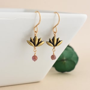 May include: A pair of gold-colored earrings featuring lotus flower pendants. Each earring has a small, faceted, pinkish-purple bead dangling below the lotus. The earrings are displayed against a white background with a green leaf visible in the background.