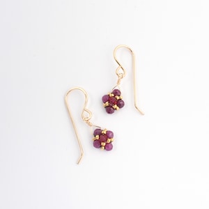 May include: A pair of gold earrings with small, red gemstone clusters. The earrings have a simple, delicate design.