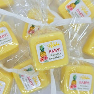 May include: A collection of square yellow soap bars individually wrapped in clear plastic with white ribbon ties. Each bar features a square label with a pineapple illustration, pink hibiscus flowers, and the text Aloha BABY! Amanda's Baby Shower.