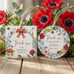 Könnte beinhalten: Zwei dekorative Anhänger mit Blumen- und Insektenillustrationen. Einer ist quadratisch mit einer roten Schleife und dem Text "Danke, dass Sie mit uns feiern!". Der andere ist rund und sagt "Danke, dass Sie Charlotte mit Liebe beschenken."