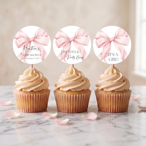 May include: Three cupcakes topped with circular paper toppers featuring a light pink bow illustration. The first topper reads Heather's BABY SHOWER September 13, 2027, the second reads TINY TOES & Pretty Bows, and the third reads IT'S A GIRL!