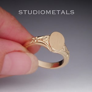Op de afbeelding: Een gouden zegelring met een ovaal vlak en gedetailleerde gravures langs de ringband. De tekst STUDIOMETALS is zichtbaar boven de ring. De ring heeft een gepolijste afwerking en een glad, plat ovaal oppervlak in het midden van de band.