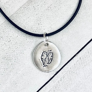 May include: A silver pendant necklace featuring a stamped design of two otters. The pendant hangs from a black cord. The otters are depicted in a simple, stylized manner, with a textured, rustic finish.