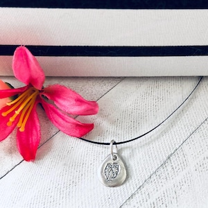 May include: A silver-toned pendant shaped like a rounded teardrop featuring an engraved design of two small figures, suspended from a thin black cord. A vibrant pink flower with yellow stamens is positioned next to the silver pendant.