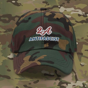Pode incluir: Um boné de beisebol camuflado com o texto "2A ANTIFASCIST" bordado na frente em fio vermelho, branco e azul.