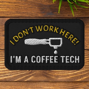 Puede incluir: Parche bordado negro con texto amarillo que dice "¡No trabajo aquí! Soy un técnico de café". Una silueta blanca de un portafiltro de máquina de café se encuentra en el centro del parche.